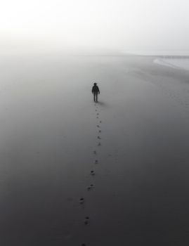 A lone figure walks along a misty beach, leaving footprints behind in the dense fog.