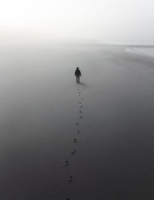 A lone figure walks along a misty beach, leaving footprints behind in the dense fog.