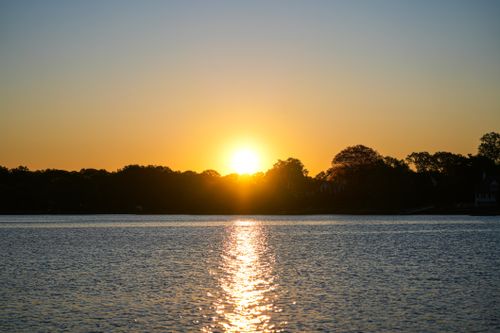 Free Tranquil sunset view with sparkling reflections over a peaceful lake, surrounded by silhouetted trees. Stock Photo