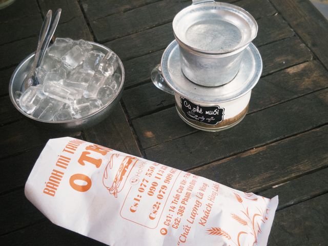 Vietnamese drip coffee beside a wrapped bánh mì on a wooden table with ice.