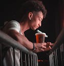 Side View Photo of Man in White T-shirt Leaning on Metal Railing Holding a Drink While Using His Phone