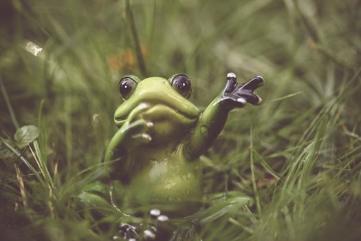 A cute ceramic frog with raised hand, surrounded by verdant grass. Perfect for whimsical decor themes.