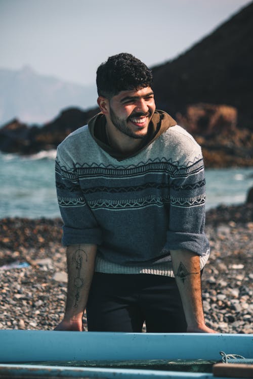 Free Young tattooed happy ethnic male in sweater standing leaned forward holding boat on pebble sea coast behind mountains and looking away on sunny day Stock Photo