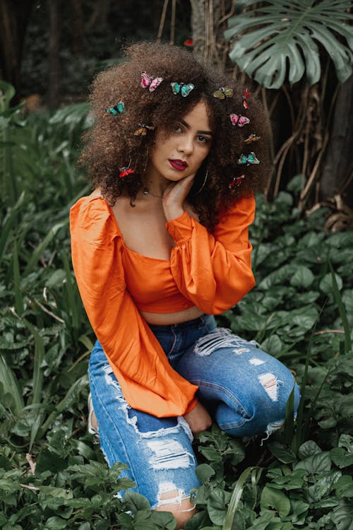 Photo of Woman in Orange Top and Blue Denim Jeans Kneeling on Green Plants Posing