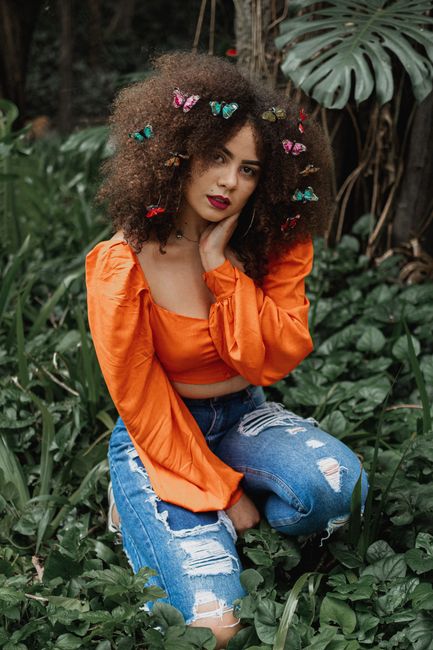 Woman in vibrant orange top posing amongst green plants with butterfly hair clips.