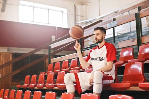 Free Low angle of young male sportsman in professional uniform sitting on sports seat of basketball court and spinning ball while looking away Stock Photo