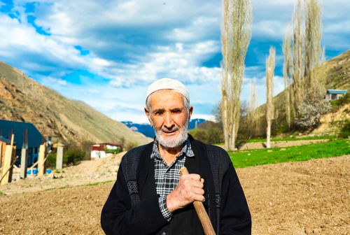Free Portrait of an elderly farmer holding a hoe against a backdrop of hills and fields under a blue sky. Stock Photo