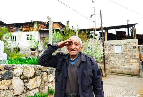 Free An elderly man offers a salute in front of rustic village houses, exuding charm and nostalgia. Stock Photo