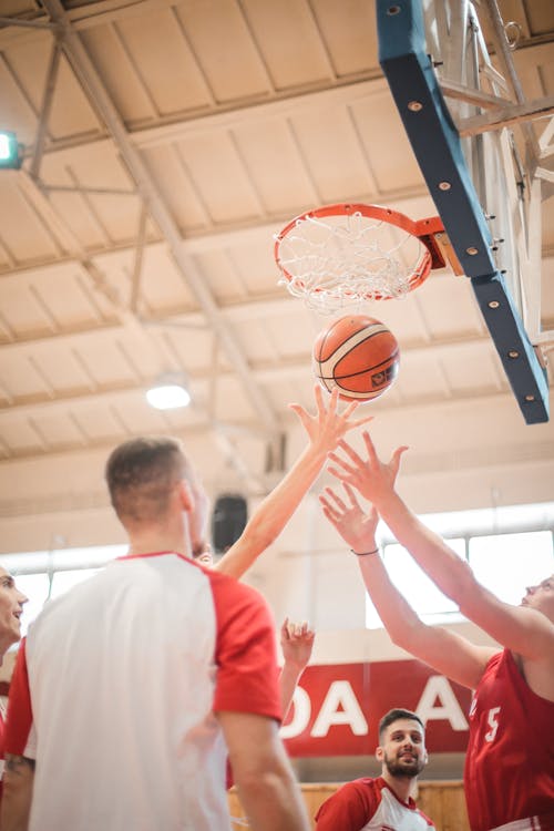 Free From below adult men in red and white sportswear catching ball from basket while playing basketball on sports arena in light contemporary sports club Stock Photo