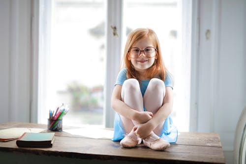 Free Joyful red haired schoolgirl in blue dress and ballet shoes smiling at camera while sitting on rustic wooden table hugging knees beside school supplies against big window at home Stock Photo