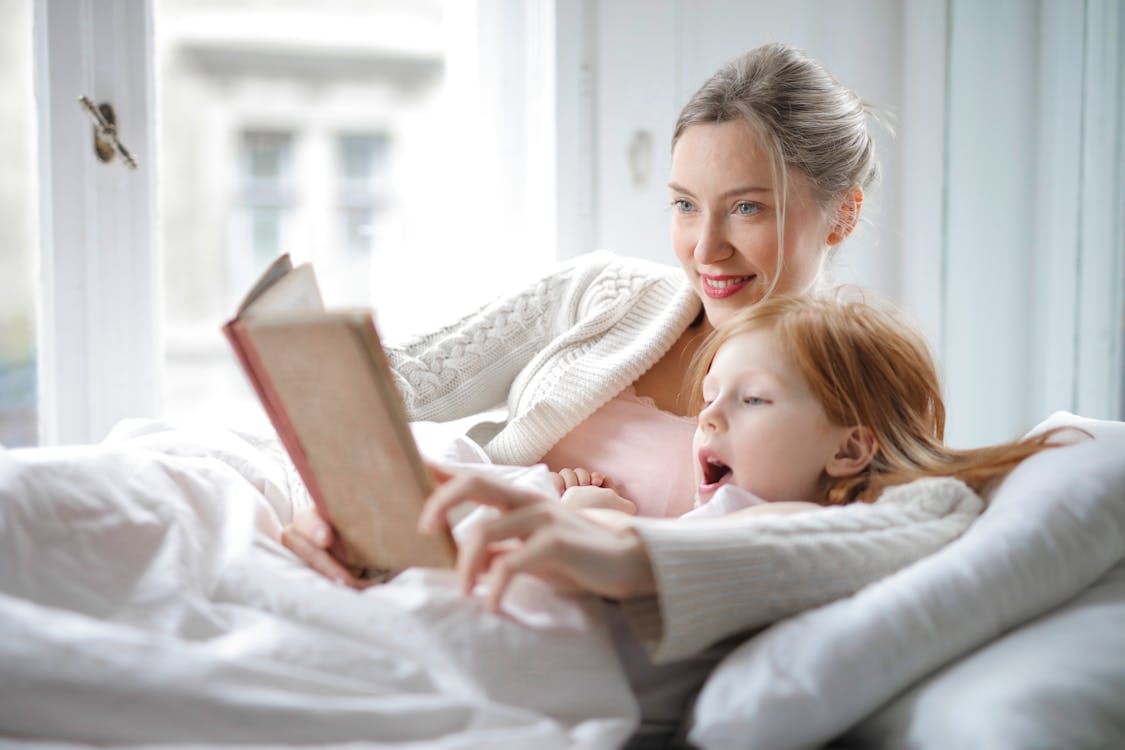 Cheerful young woman hugging cute little girl and reading book together while lying in soft bed in light bedroom at home in daytime