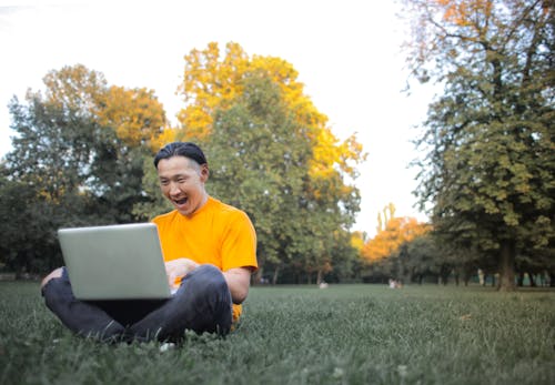 黃船員的脖子t恤和灰色的褲子，坐在綠草地上，使用筆記本電腦的人