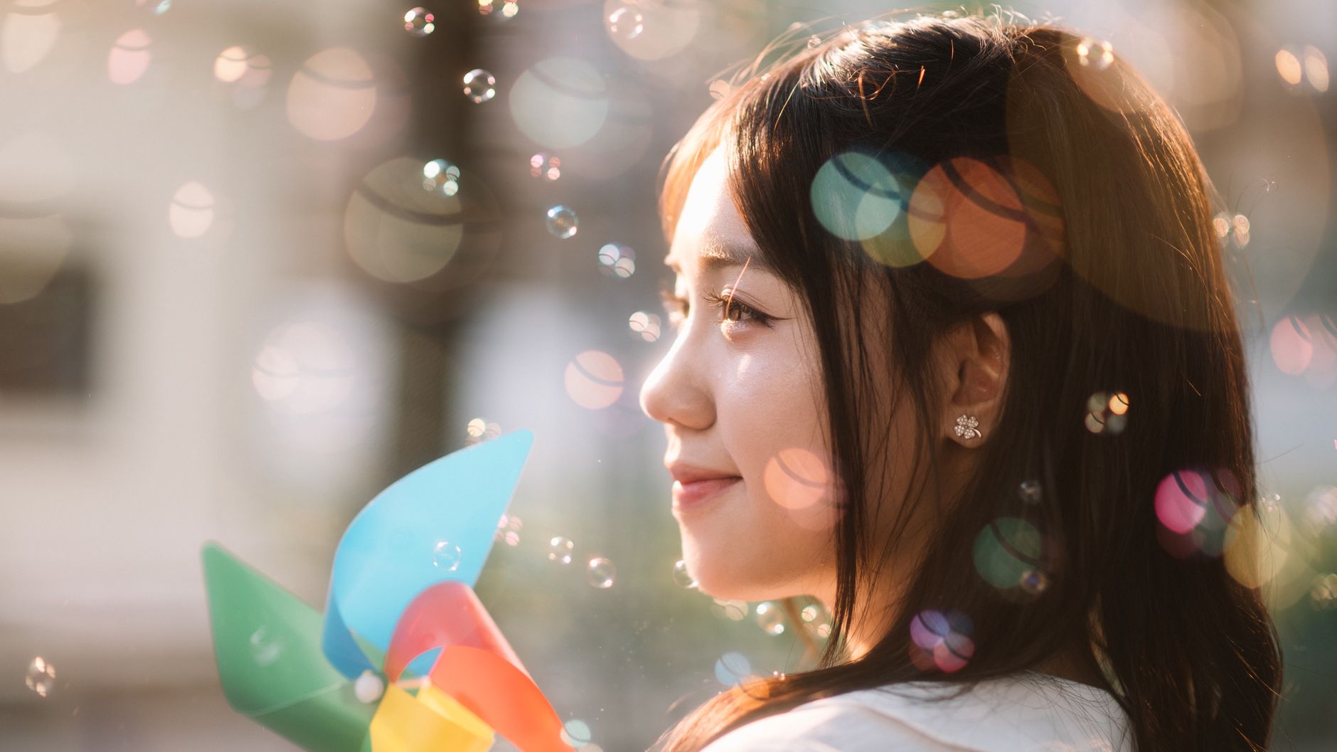 A smiling young woman holds a colorful pinwheel amidst floating bubbles outdoors.