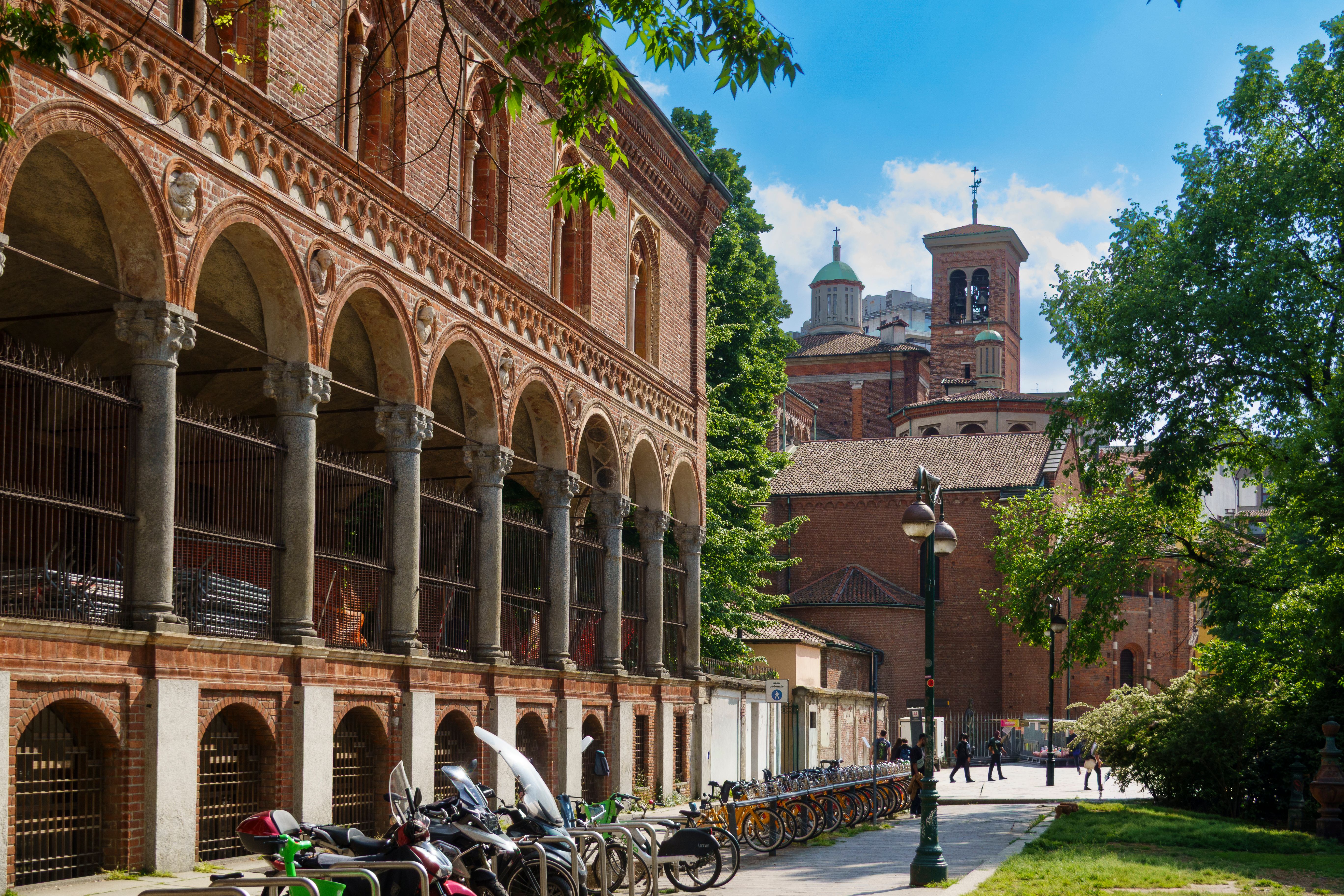 Igreja de Santa Maria delle Grazie em Milão, Itália, com arcos de tijolos vermelhos, jardins frondosos e pedestres sob sol forte