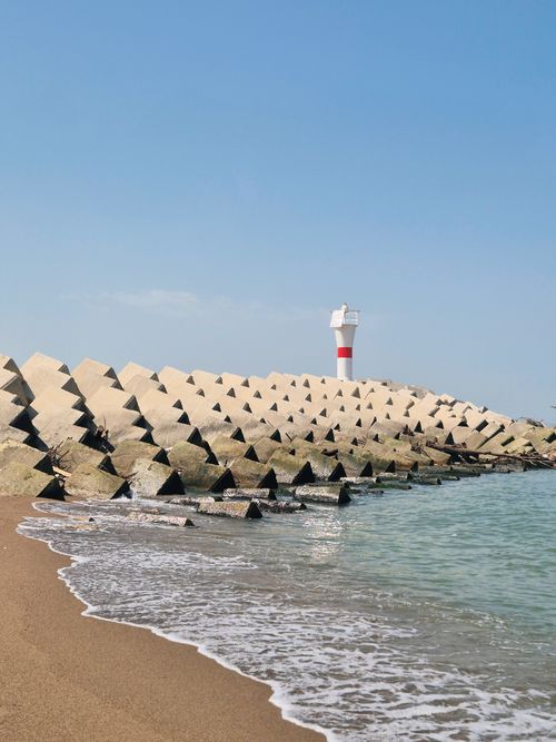 Free A serene coastal scene featuring a lighthouse and concrete breakwater under a clear blue sky. Stock Photo