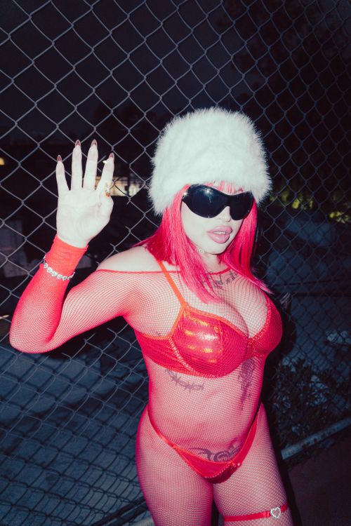 Free Edgy style portrait of a woman in red fishnet attire, sunglasses, and a white fur hat at night. Stock Photo