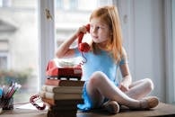 Low angle of calm redhead preteen lady in blue dress and beige sandals looking away and having phone call using retro disk telephone on stack of books while sitting with legs crossed on wooden table against window at home