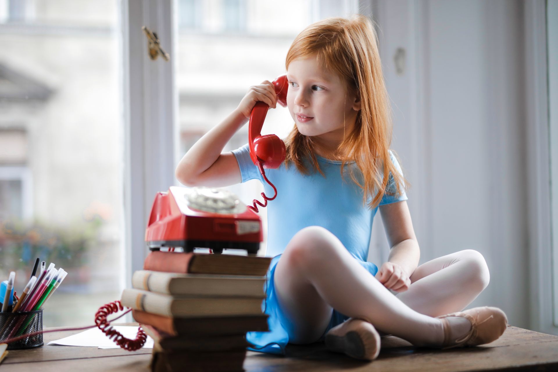 Low angle of calm redhead preteen lady in blue dress and beige sandals looking away and having phone call using retro disk telephone on stack of books while sitting with legs crossed on wooden table against window at home