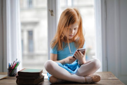 Free Red haired charming schoolgirl in blue dress browsing smartphone while sitting on rustic wooden table with legs crossed beside books against big window at home Stock Photo