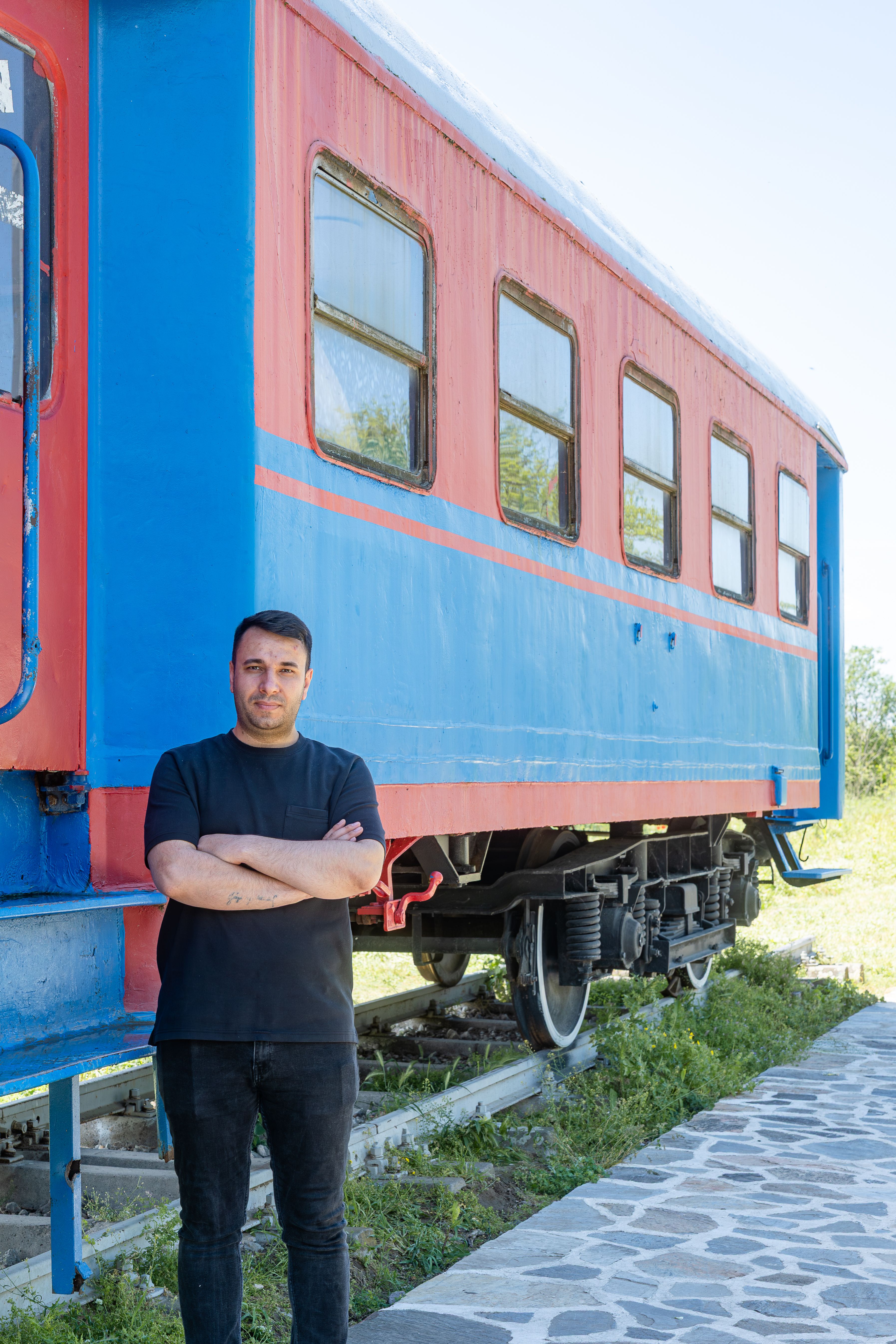 A man stands beside a vintage red and blue train car outdoors on a sunny day.