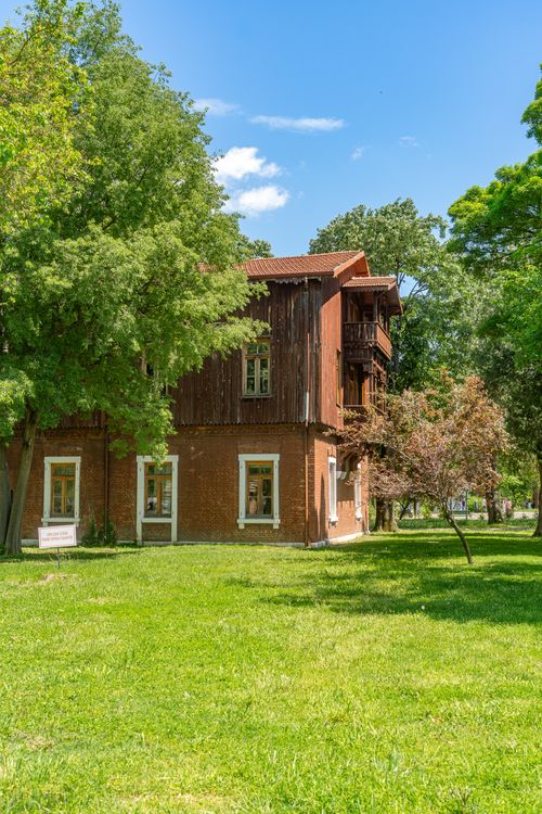 Free A charming rustic house nestled among lush trees and a vibrant green lawn under a clear blue sky. Stock Photo