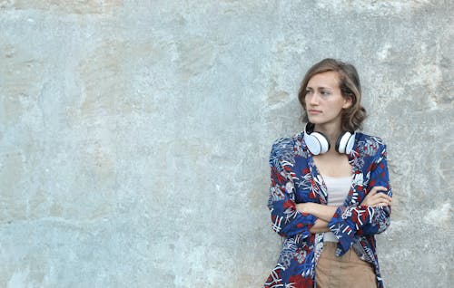 Foto Di Donna In Camicia A Maniche Lunghe Floreale Blu E Bianca Con Le Cuffie Sul Collo Appoggiato Sul Muro Grigio Che Osserva Via