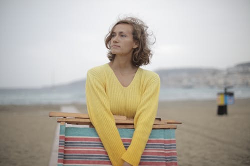 Calm female tourist with folded deckchair standing alone on seashore in overcast weather