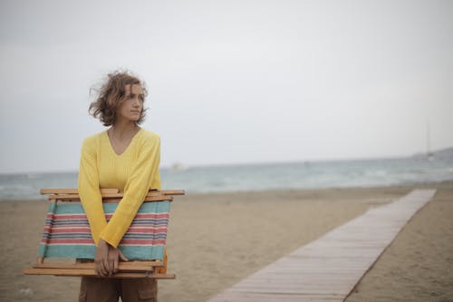 無料 木製の折りたたみ式ビーチチェアを運ぶビーチに立っている黄色の長袖シャツの女性の写真 写真素材