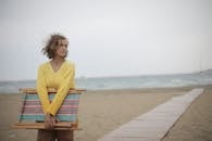 Photo of Woman in Yellow Long Sleeve Shirt Standing at the Beach Carrying Wooden Folding Beach Chair