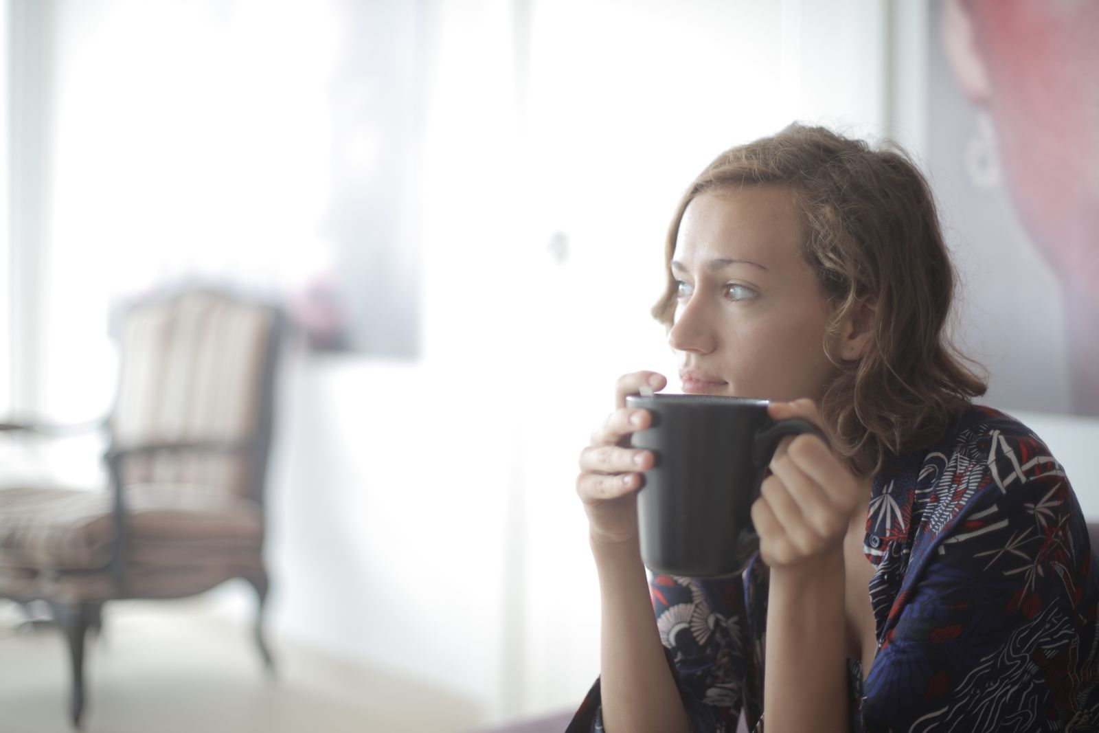 A relaxed woman in a floral shirt enjoys a hot drink while sitting in a bright, cozy room.