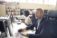 High angle of pensive shocked businessman in elegant clothes focusing on screen and interacting with computer while sitting at table near African American female coworker in light spacious office