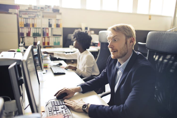 Adult Surprised Executive Man Using Computer Beside Ethnic Colleague In Open Space Office