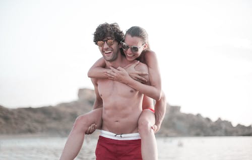 Foto De Un Hombre En Topless En Pantalones Cortos Rojos Con Una Mujer En La Espalda Mientras Está En La Playa