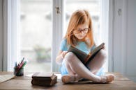 一位可爱的年轻女孩戴着眼镜，在室内靠窗读书，画面捕捉了她的纯真与专注