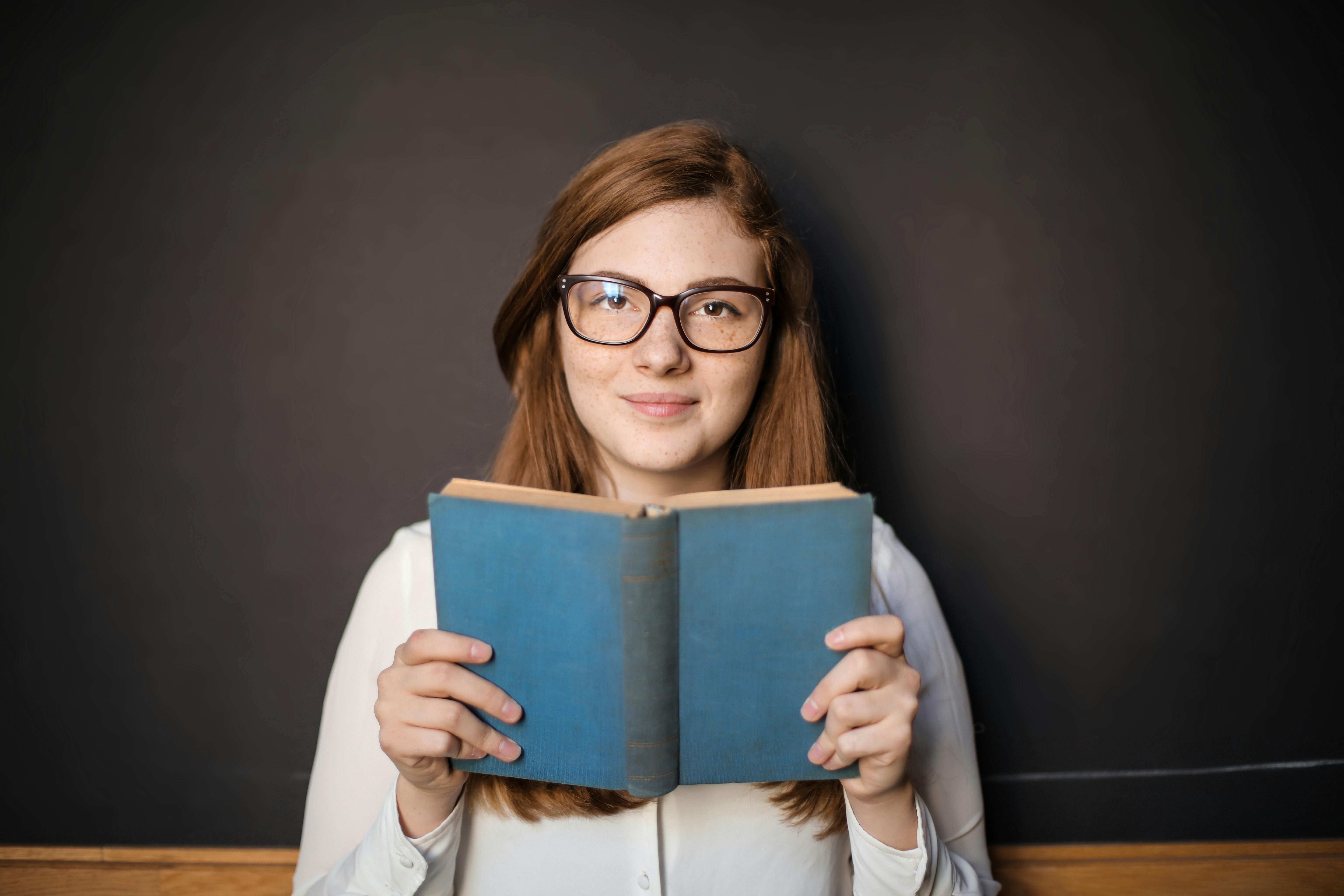 woman in white long sleeve shirt wearing eyeglasses holding blue book