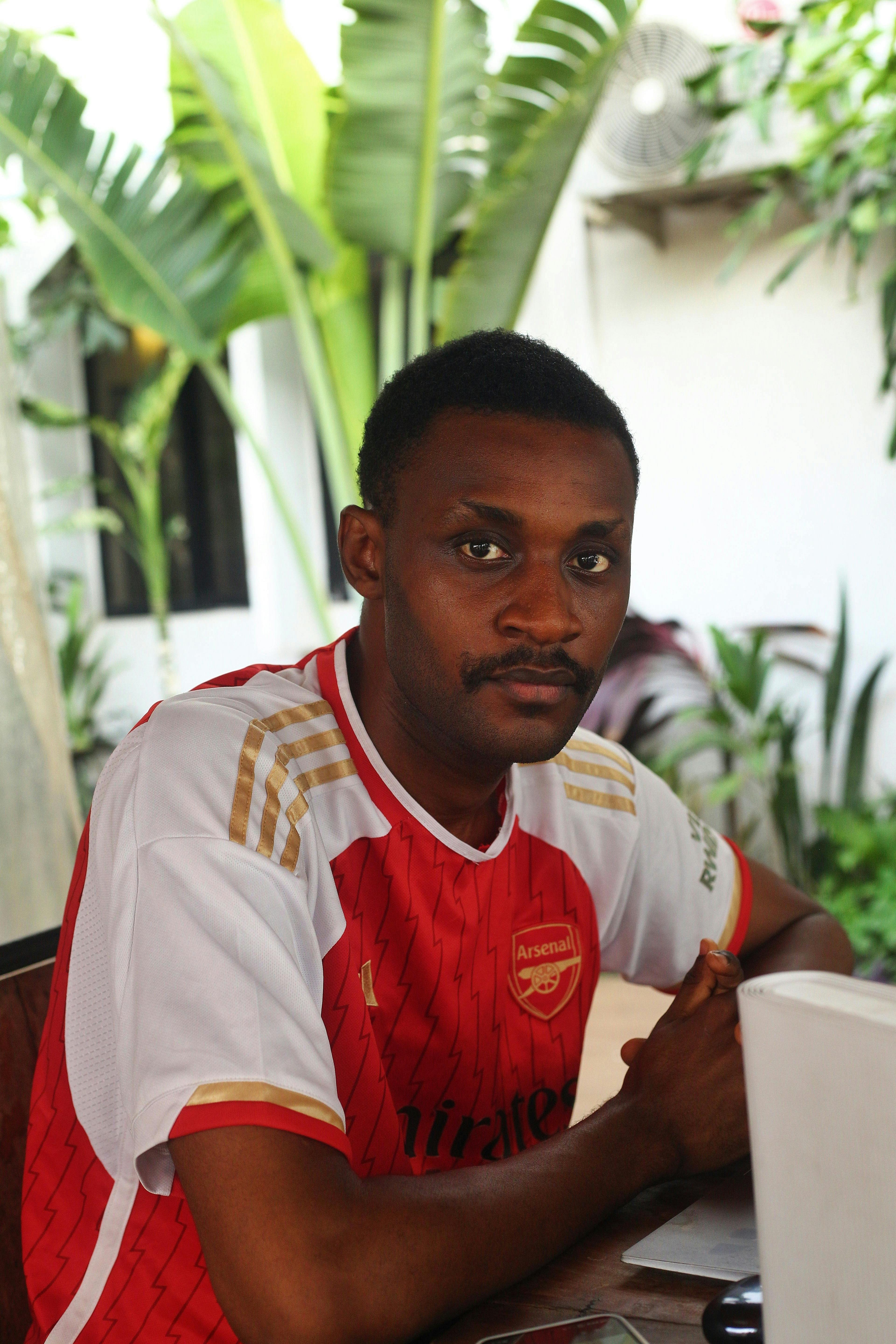 Man wearing a sports jersey sitting outdoors with a laptop, surrounded by tropical plants.
