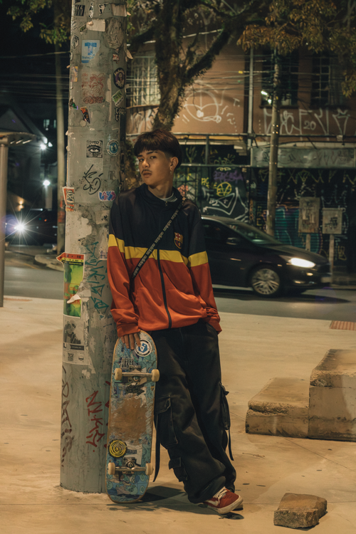 Free Teenage skater in colorful jacket and skateboard leaning on graffiti-covered pole at night city street. Stock Photo