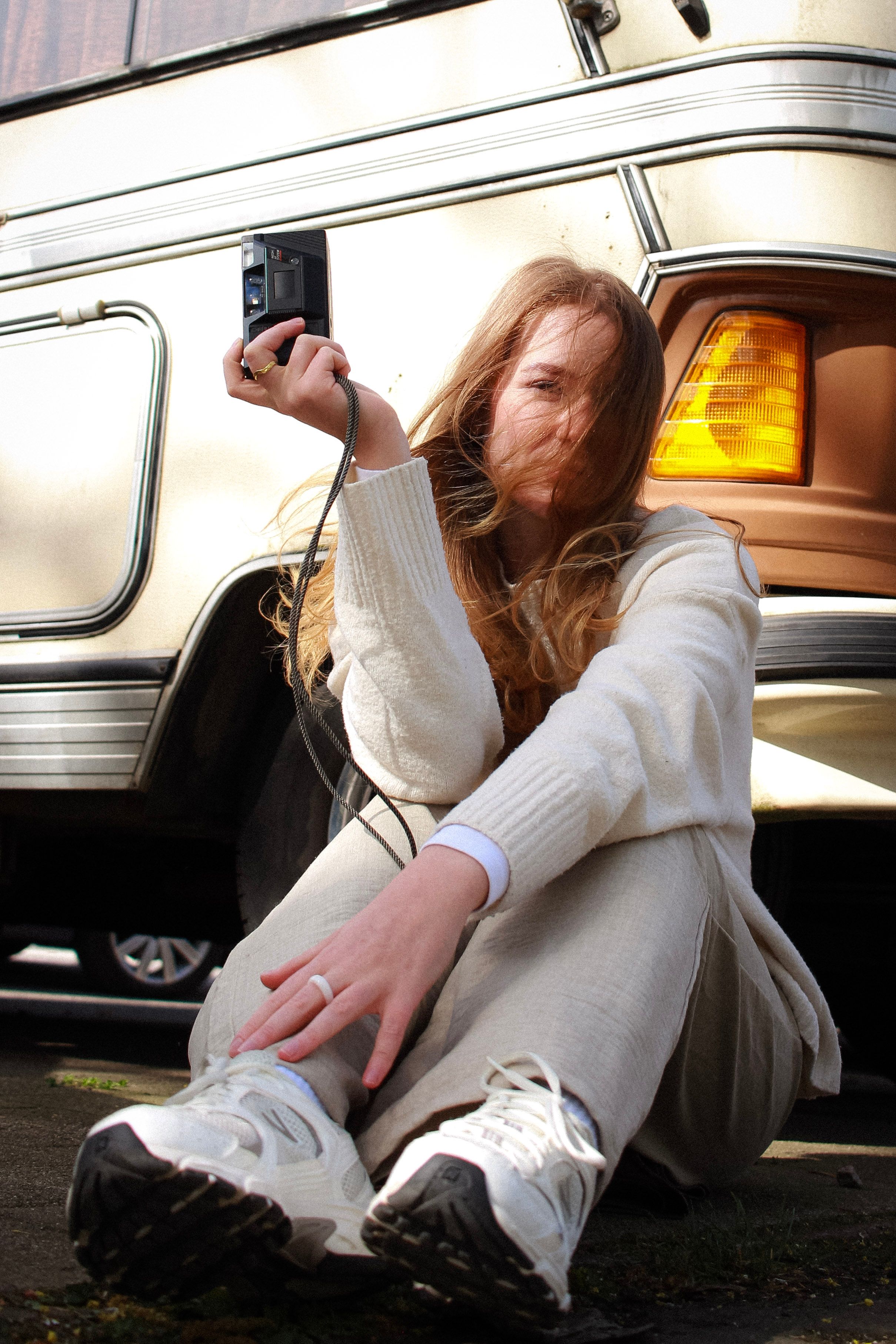 A woman with blonde hair sitting outside, holding a camera, next to a vehicle in Hamburg.