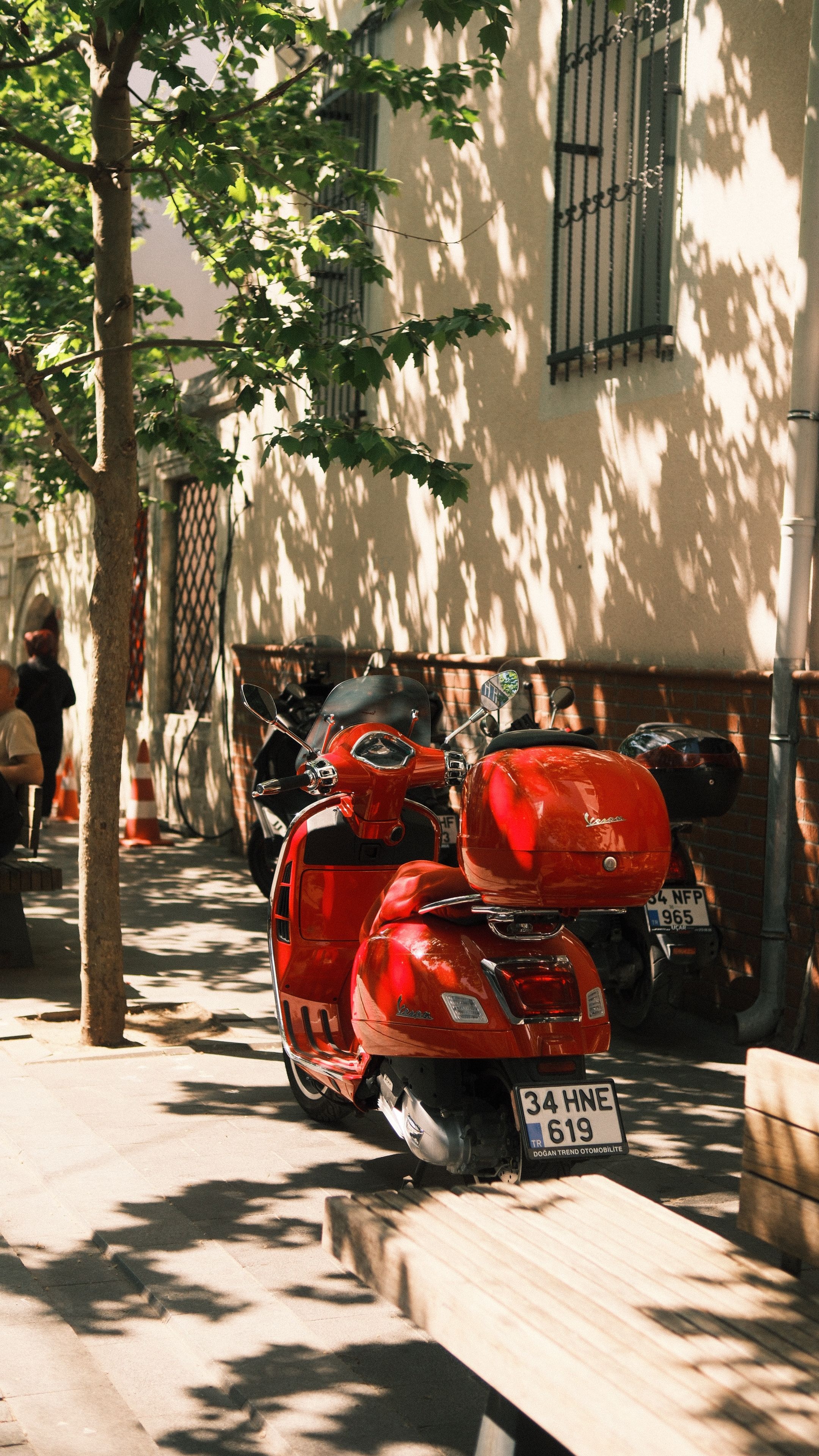 A vibrant red Vespa scooter parked on a tree-lined street during the day.