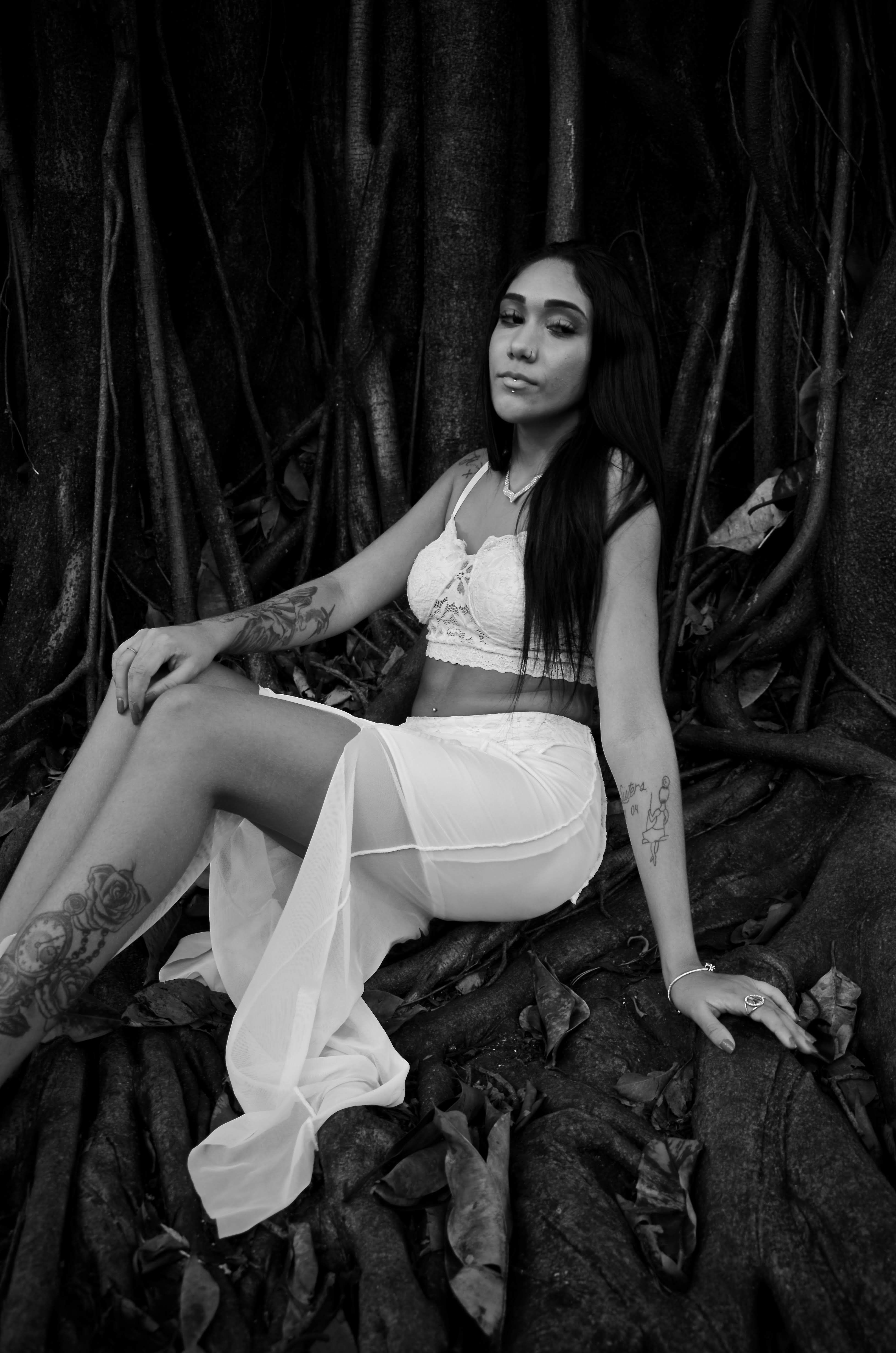 Black and white portrait of a tattooed woman sitting gracefully among tree roots.