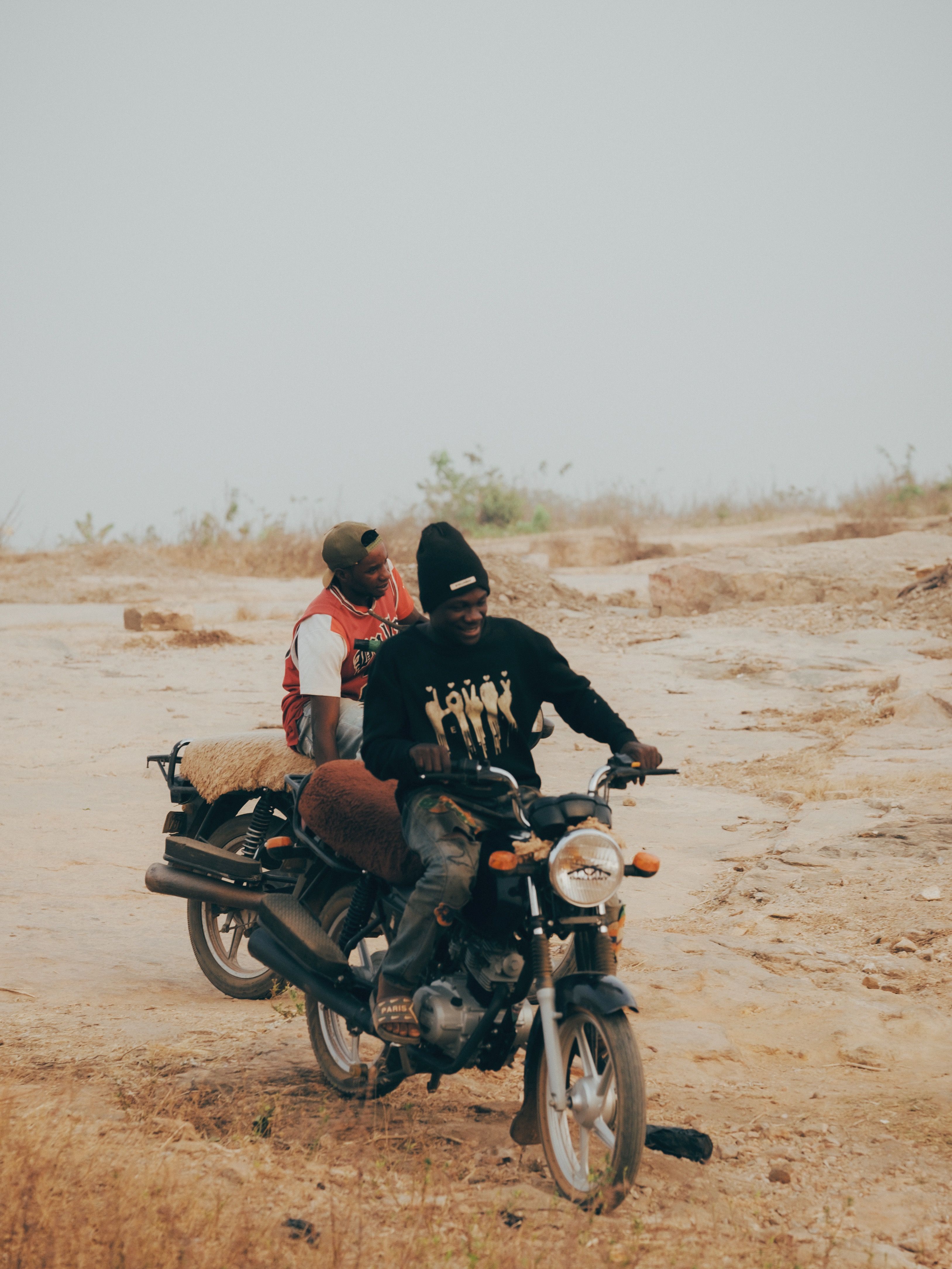 Two men ride a motorcycle on a rural dirt path surrounded by dry landscape.