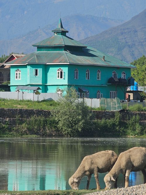 Free A beautiful green building by a lake with grazing sheep, set against a mountainous backdrop. Stock Photo