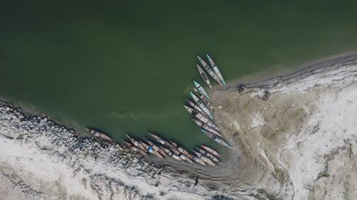 Free Aerial shot of traditional wooden boats moored along a riverbank in Bangladesh. Green water and rocky shore create a serene landscape. Stock Photo