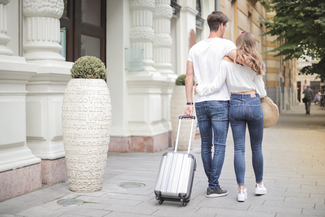 Free Back view of traveling couple in love wearing casual clothes walking with luggage and hugging while strolling along sidewalk together during vacation Stock Photo