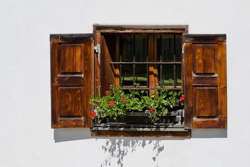 Free A rustic window with wooden shutters and vibrant flowers adds charm to a white wall. Stock Photo