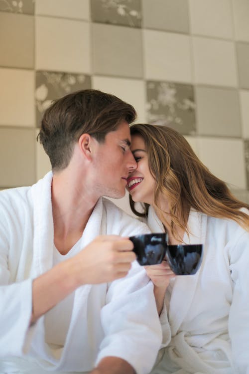 Free Young gentle couple wearing bathrobes sitting close to each other with cups of hot drink and hugging with closed eyes at home Stock Photo