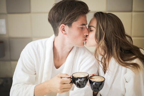 Gratis Foto Pasangan Dengan Jubah Mandi Putih Berciuman Saat Memegang Cangkir Kopi Keramik Hitam Foto Stok