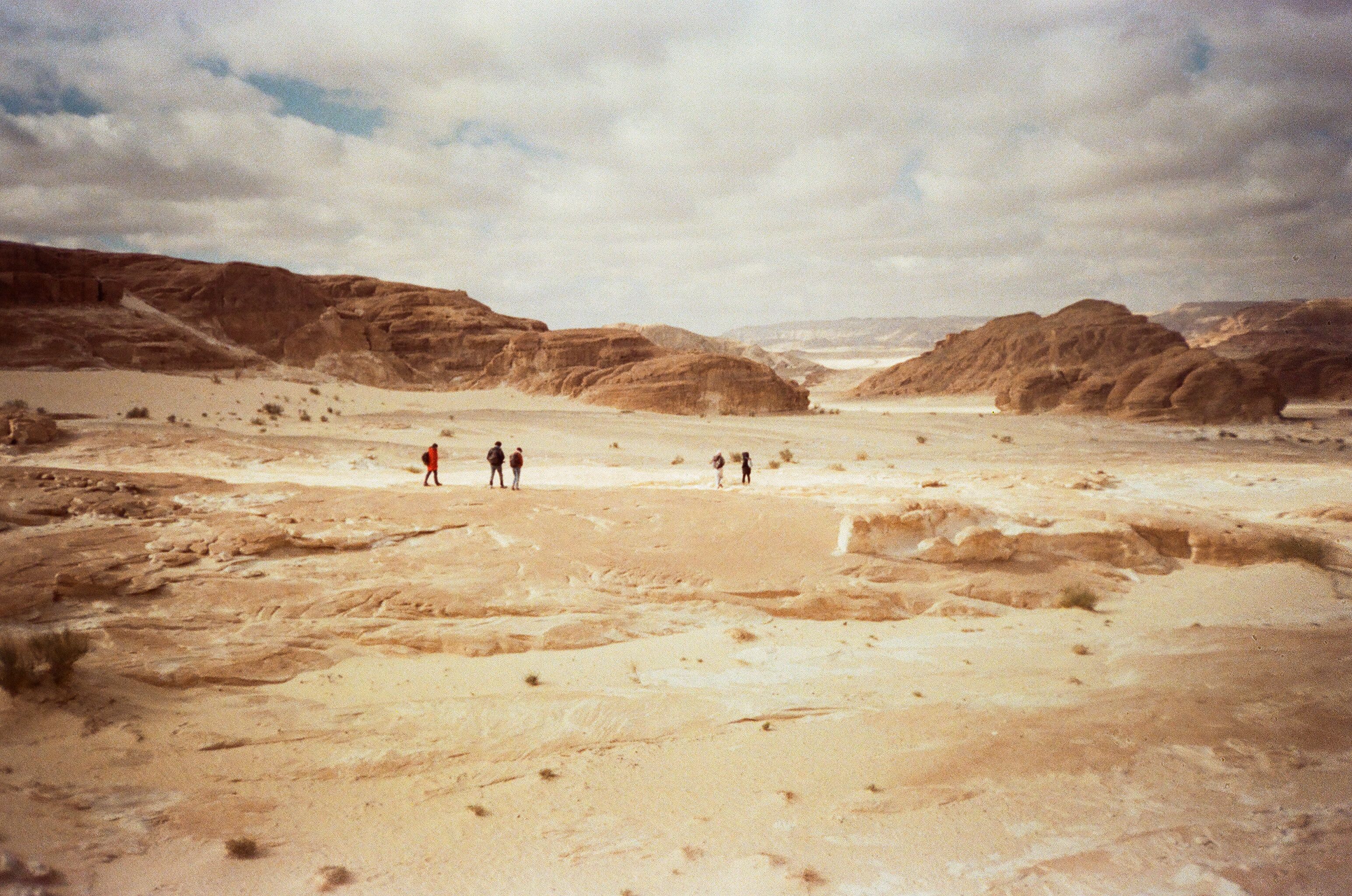 Group of hikers traversing a vast desert with rocky formations under a cloudy sky.