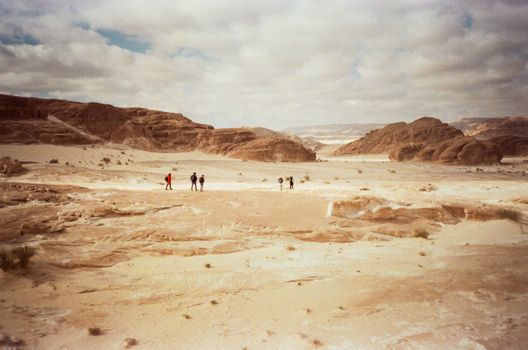 Group of hikers traversing a vast desert with rocky formations under a cloudy sky.