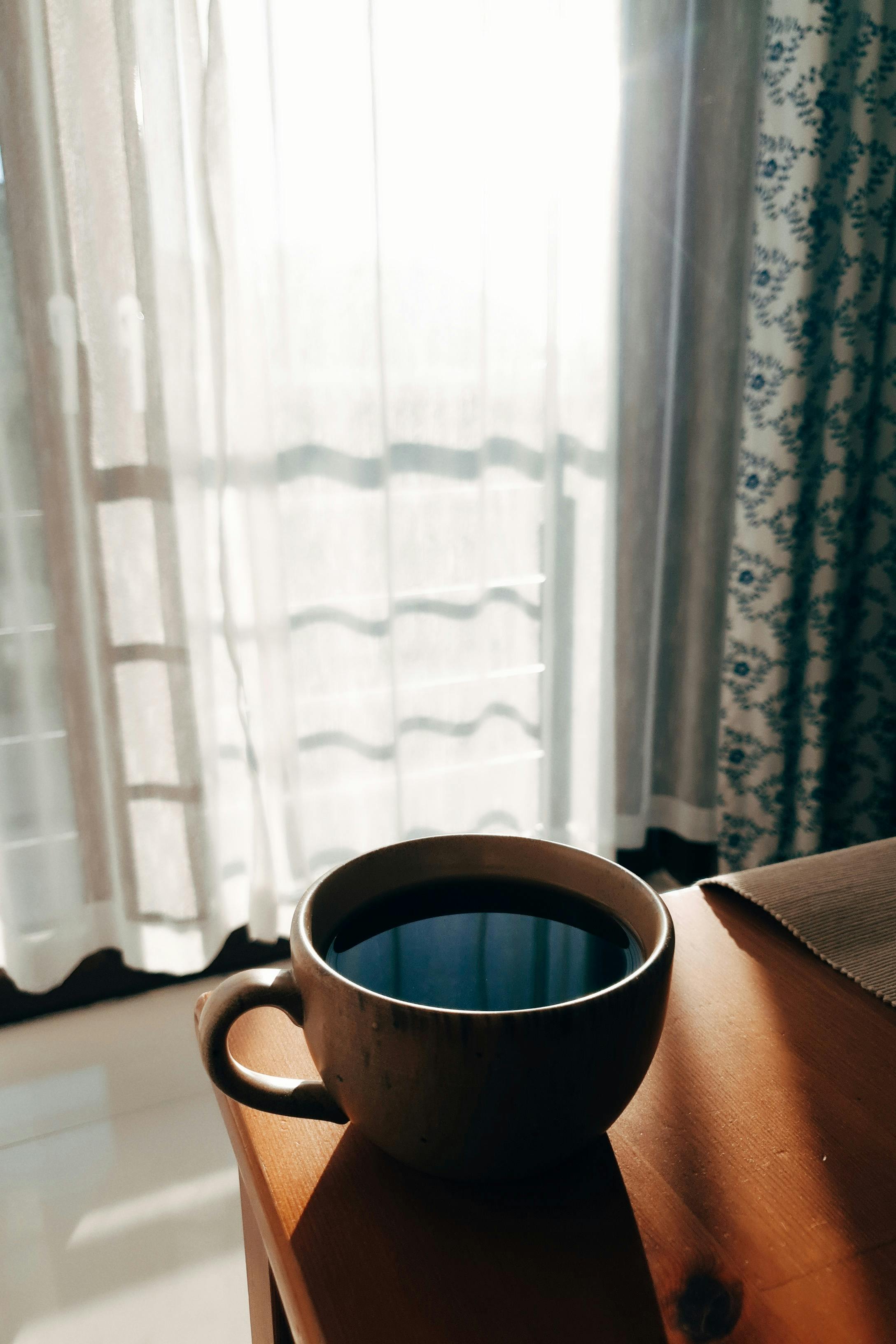 Warm morning light streaming through curtains onto a table with a cup of coffee.